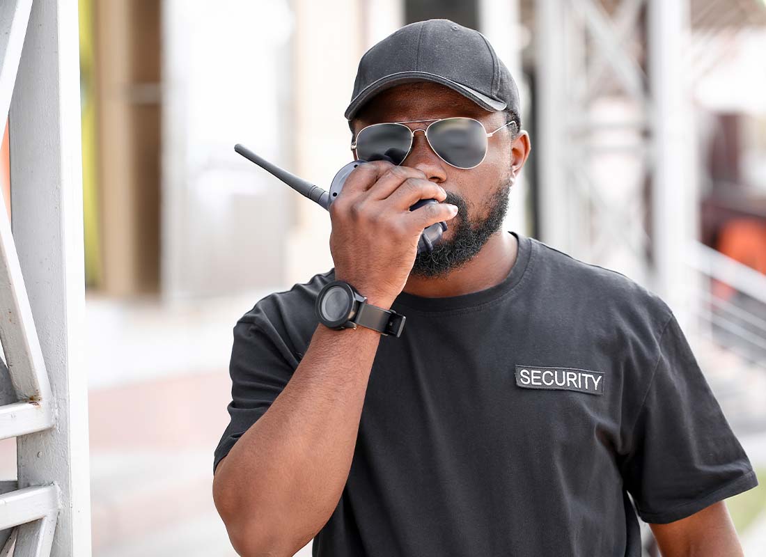 Security Guard Insurance - Security Guard Using a Walkie Talkie and Communicating Security Concerns With Head Office Outdoors in a Baseball Cap and T-Shirt