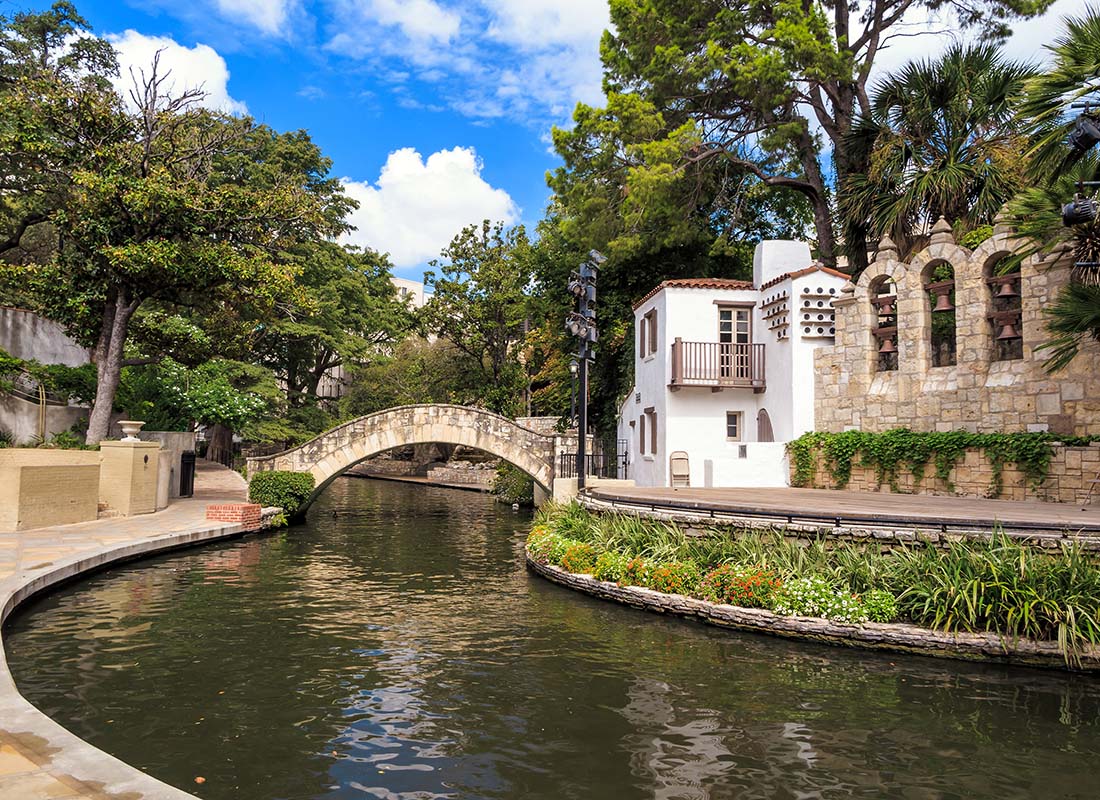 Read Our Reviews - Scenic Landscape of Historical Buildings Next to a River with a Walking Bridge Across Surrounded by Green Foliage in San Antonio Texas